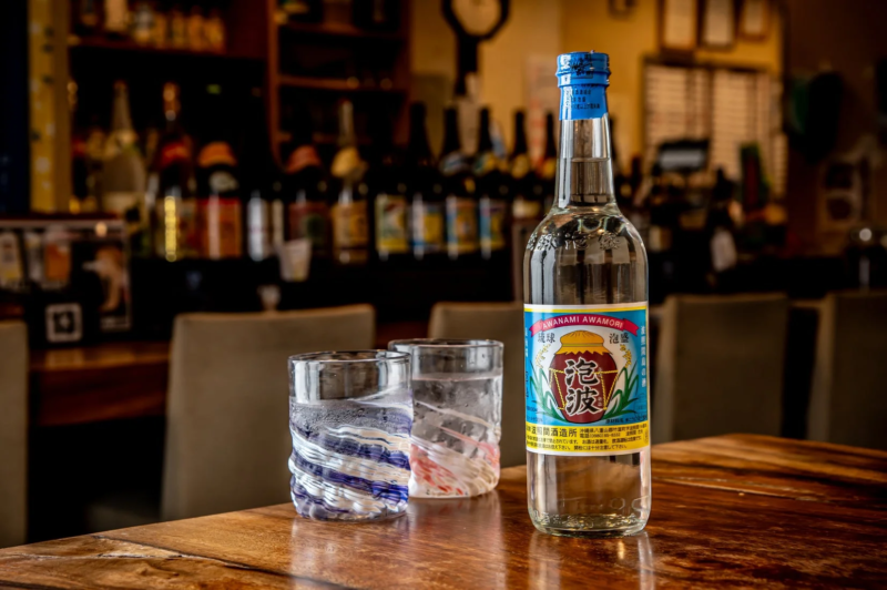 木製のテーブルに沖縄の伝統的なお酒である泡盛「泡波」のボトルと、水が入った二つの琉球ガラスのグラスが置かれています。背景には多くの酒瓶が並べられており、居酒屋のような落ち着いた雰囲気が漂っています。