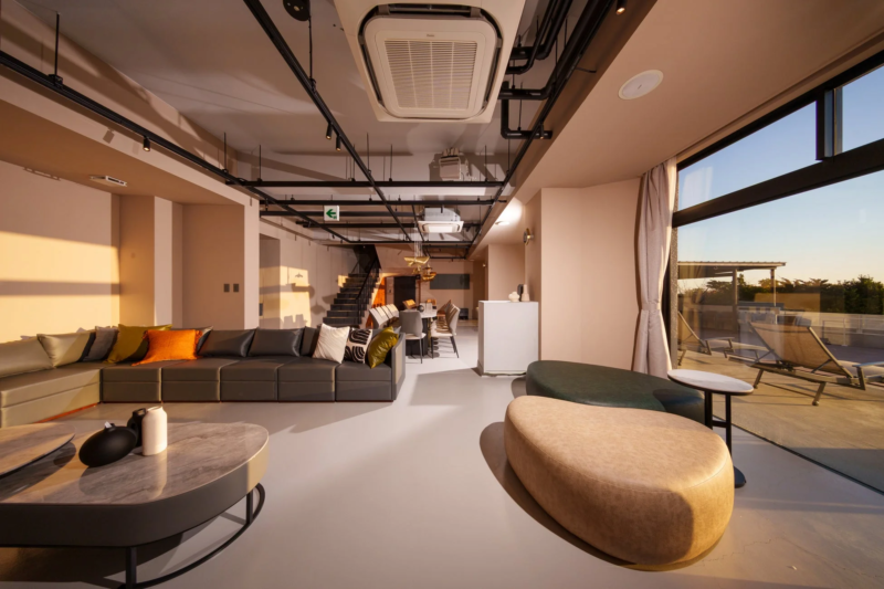 広々としたモダンなラウンジ空間で、大きな窓からは明るい日差しが差し込み、屋外のテラスが見えます。快適なソファとスタイリッシュな家具が配置され、落ち着いた雰囲気です。