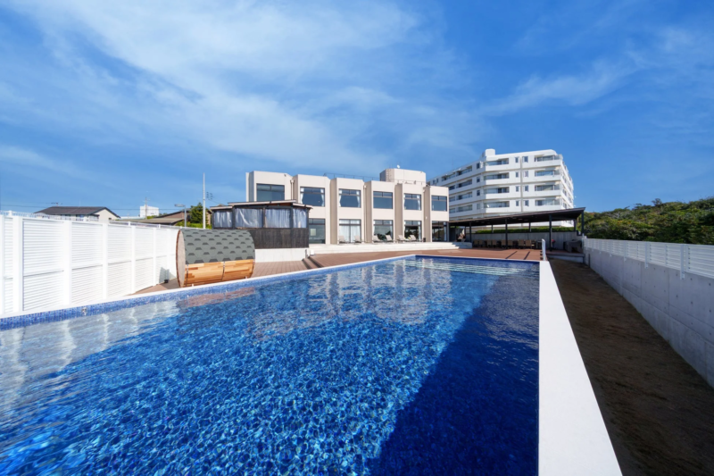 青い空の下、広々とした屋外プールとユニークなバレルサウナが特徴的なリゾート施設の一角。モダンな建物に囲まれ、開放感あふれる空間でリラックスできる様子が伺える。