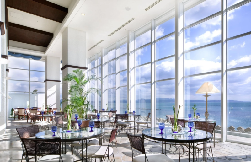 広々とした空間に大きな窓からオーシャンビューが広がる、明るく開放的なレストランまたはラウンジの風景です。ガラスのテーブルと籐の椅子が並び、青いグラスや観葉植物が配置されています。