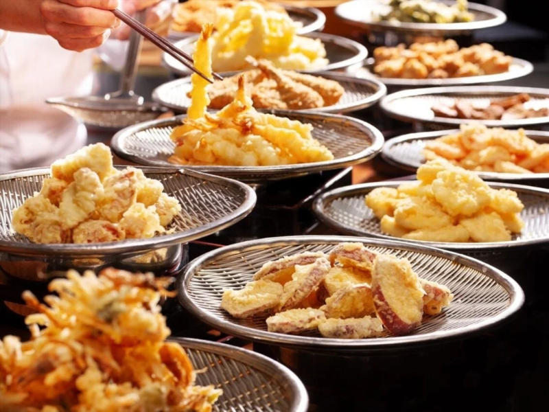 揚げたての様々な天ぷらが多数の網皿に並べられ、食欲をそそる光景です。手元には箸で海老天をつまむ様子が捉えられており、種類豊富な日本食の美味しさが伝わります。