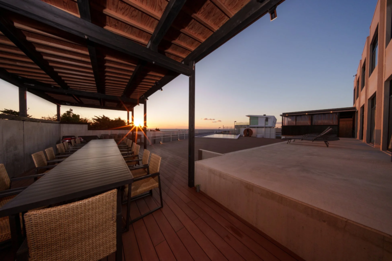 夕焼けが美しい広々とした屋外テラスの風景。木製のデッキにはパーゴラの下に長いダイニングテーブルと椅子が並び、遠くには海と小さなプール、そしてモダンな建物が見える。リラックスできる豪華な空間が夕暮れ時に映し出されている。
