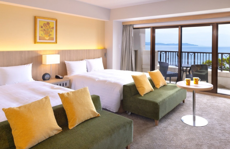海が見えるバルコニー付きのホテル客室です。ツインベッドとソファが配置され、明るく開放的な空間が広がっています。窓の外には広大な海が望め、リゾート滞在にぴったりのリラックスできる雰囲気です。