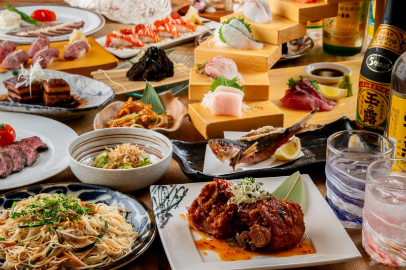 刺身、寿司、肉料理、魚料理、麺類など、多種多様な和食が豪華に並べられた食卓の風景。沖縄の泡盛も添えられており、賑やかな宴の雰囲気を伝えています。
