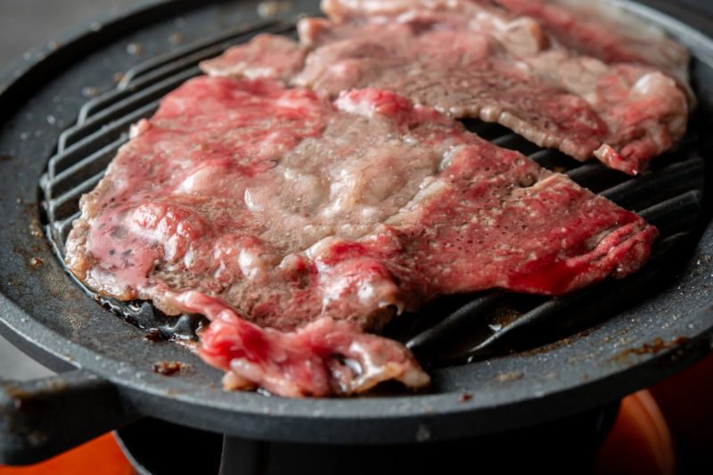 鉄板のグリルで焼かれている薄切りの牛肉のクローズアップ画像です。肉はまだ生の部分が多く、焼ける途中の様子が分かります。脂と赤身のバランスが良い霜降り肉で、食欲をそそる一品です。