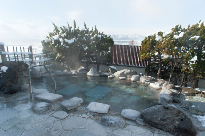雪が積もった冬の露天風呂の風景。湯気が立ち上る温泉が広がり、周囲は岩や雪をかぶった木々に囲まれている。遠くには雪山と街並みが見える。