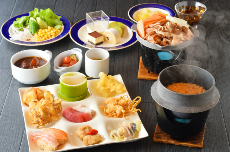 様々な種類の料理が並べられた豪華な食事の風景です。釜飯や鍋物、刺身、寿司、天ぷらといった和食の他、サラダ、デザート、シチューのような洋食も含まれており、旅館やホテルの会席料理を思わせます。