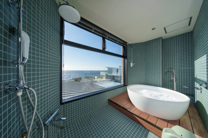 海を見渡せるモダンなバスルームです。オーシャンビューの大きな窓があり、木製デッキの上に置かれた白いバスタブとシャワーエリアが特徴的。緑のタイルが印象的な、リラックスできる空間です。