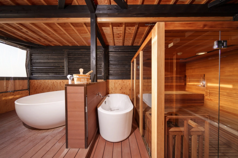 木製の壁と天井が特徴的なモダンな温浴施設で、複数の白いバスタブとガラス張りのサウナ室が配置されています。落ち着いた雰囲気でリラックスできるプライベートな空間です。