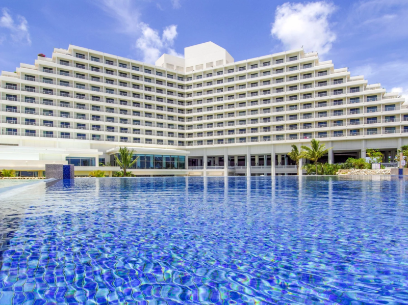 青い空と白い雲の下、広々とした屋外プールが手前に広がり、その奥にはテラスが連なる大規模な白いリゾートホテルが建っています。リゾート感あふれる景色です。