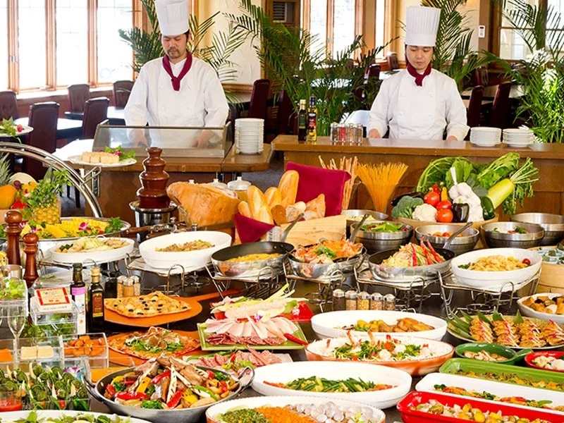 豪華なビュッフェ料理が並ぶレストランの様子。二人のシェフが準備をしており、サラダ、パスタ、肉料理、パン、デザートなど、多種多様なメニューが豊富に提供されている。