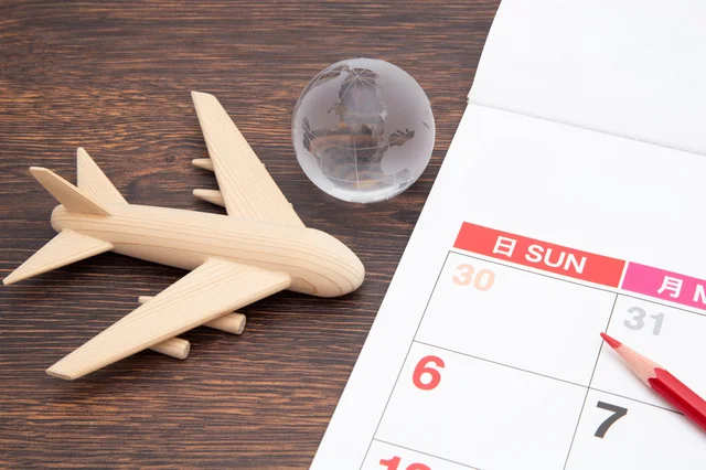 木製の飛行機模型、地球儀、カレンダー、赤鉛筆が木目のテーブルに置かれ、旅行や未来の計画を連想させる画像です。カレンダーには「日 SUN」や日付が書かれています。