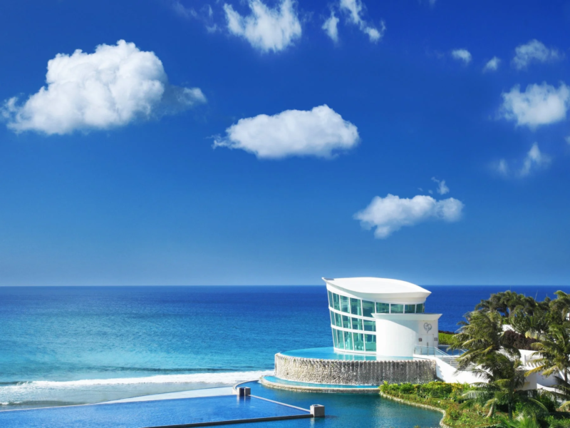 晴れた青空の下、海辺に建つモダンな白いチャペルとインフィニティプールが印象的なリゾート風景です。白い雲が浮かぶ広大な海と調和した美しい光景が広がっています。