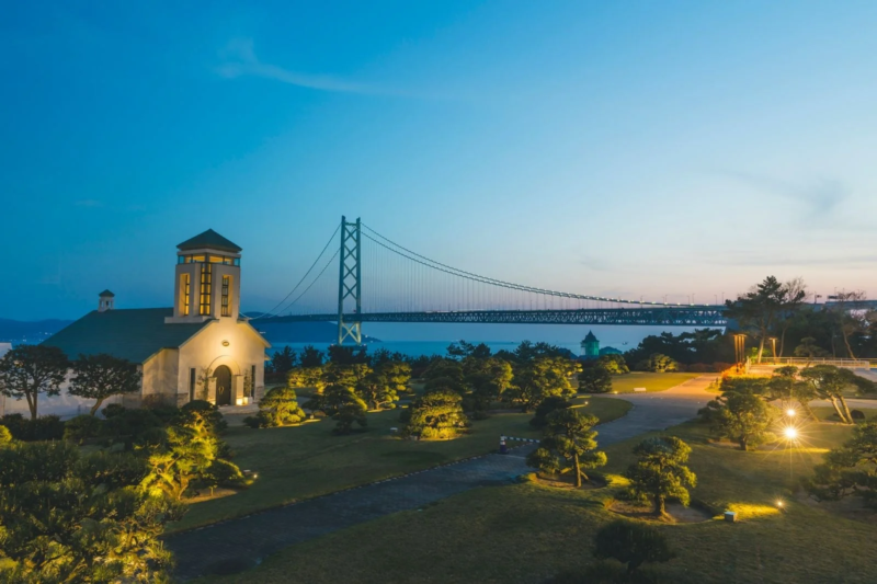 夕暮れ時、ライトアップされた美しいチャペルと手入れされた庭園が広がる風景です。背景には壮大な吊り橋が海上に架かり、幻想的な夜景を演出しています。青い空が広がり、静かでロマンチックな雰囲気が漂います。