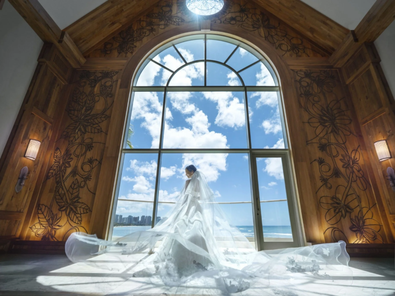 豪華な木目調のチャペルで、花嫁が大きなアーチ窓の前に立っています。窓の外には青い空と海、そして街並みが広がり、風になびくウェディングドレスとベールが美しい瞬間を捉えています。