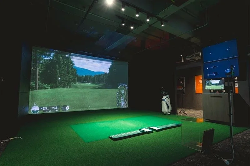 屋内のゴルフシミュレーター施設で、大きなスクリーンに美しいゴルフコースが映し出されています。人工芝の打席やゴルフバッグが設置され、リアルな環境でゴルフの練習やプレイを楽しめる様子が伝わります。