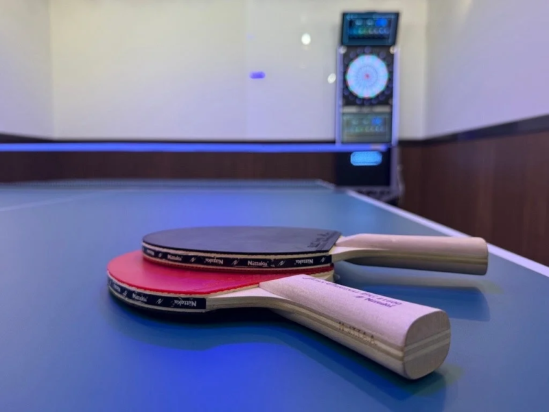 青い卓球台の上に赤い面と黒い面の卓球ラケットが2本重ねて置かれています。背景にはダーツボードが見え、スポーツやレジャーを楽しめる空間の様子が伺えます。