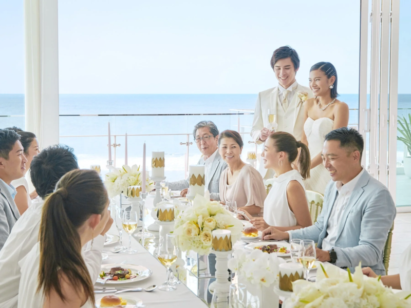 海が見える明るい会場で、新郎新婦とゲストが笑顔で食事を楽しむ結婚披露宴の様子。白い花と豪華なテーブルセッティングが祝福の雰囲気を盛り上げています。