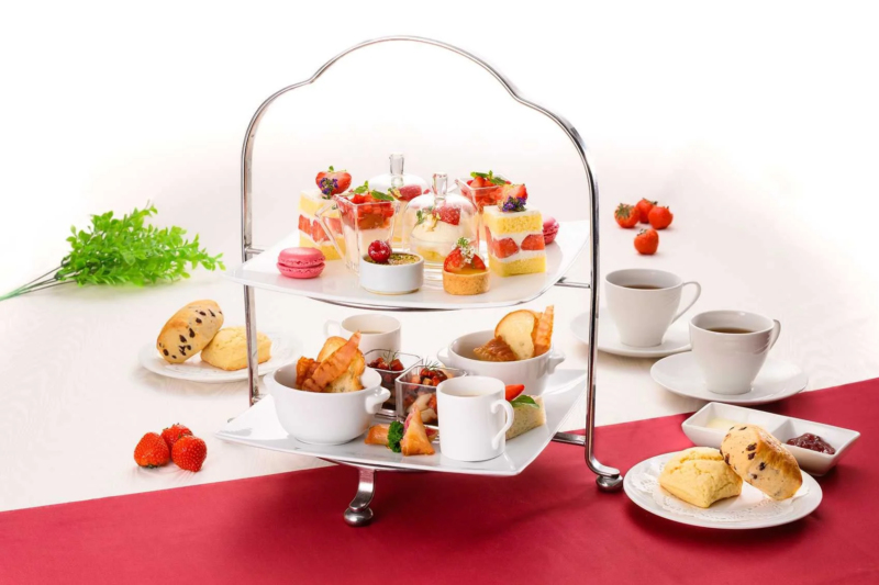 白いテーブルに赤い布が敷かれ、豪華な二段のアフタヌーンティーセットが並んでいます。いちごのケーキ、マカロン、スコーン、軽食、紅茶などが美しく盛り付けられ、優雅なティータイムを思わせる華やかな食卓です。