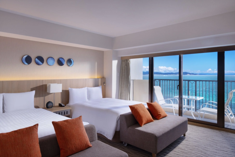 オーシャンビューの広々としたホテル客室。ツインベッドとソファが配置され、窓の外には青い海と空が広がるバルコニーがあります