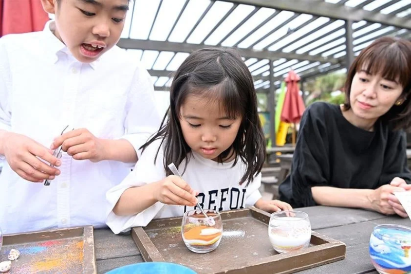 屋外のテラス席で、子供たちが母親と一緒にサンドアート体験を楽しんでいる様子。