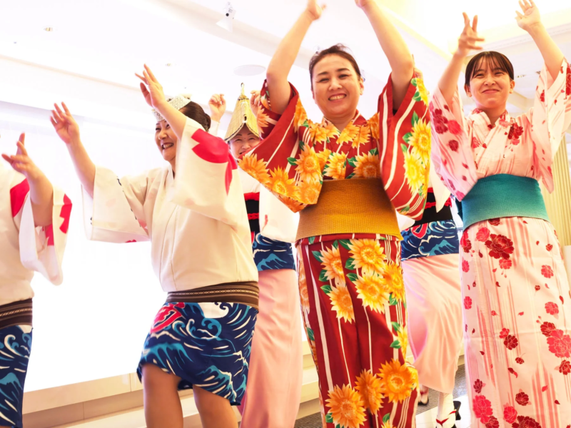 日本の伝統的な衣装を身につけた女性たちが、笑顔で両腕を上げて楽しそうに踊っています