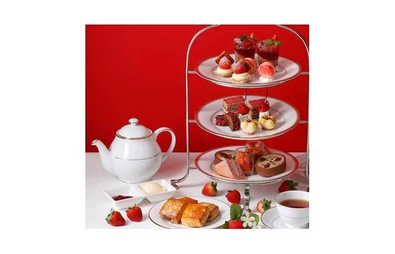赤い背景の前で、豪華なアフタヌーンティーセットが並べられています。三段スタンドには様々なスイーツやセイボリー、テーブルにはティーポット、スコーン、フレッシュないちごが添えられています。