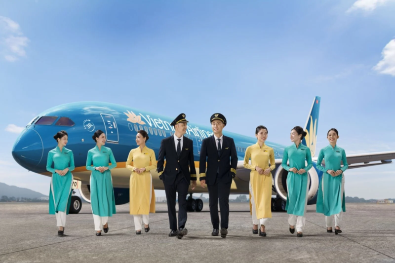 ベトナム航空のパイロット2名と客室乗務員6名が、青い機体を背景に空港の滑走路を歩いている集合写真です。