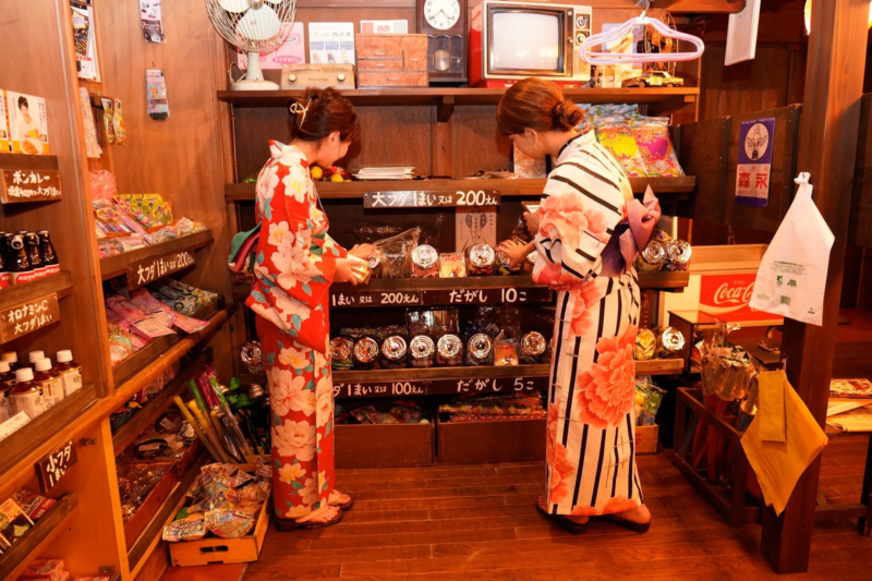 浴衣姿の二人の女性が、昭和レトロな雰囲気の駄菓子屋で買い物を楽しんでいる様子