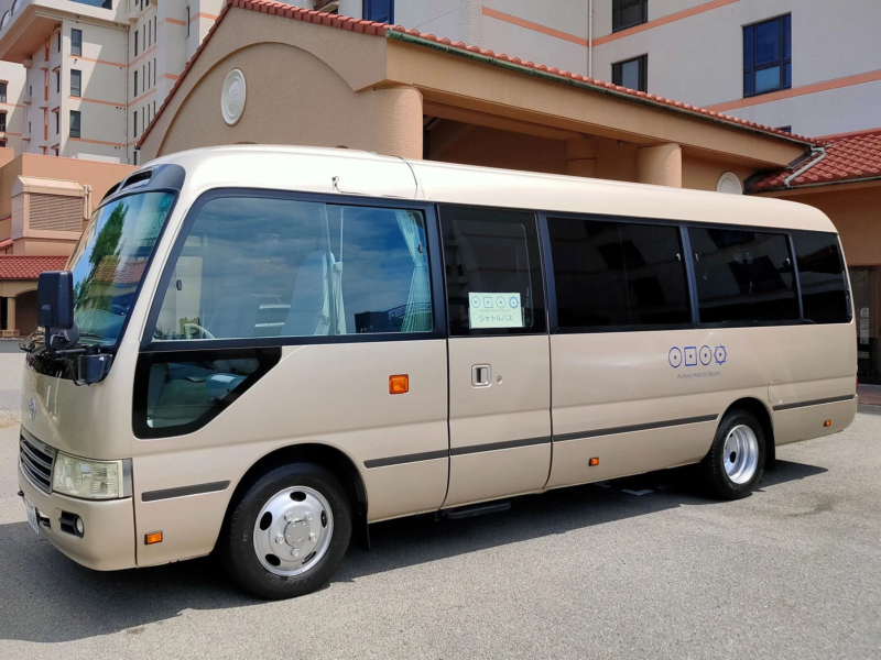 ベージュ色のマイクロバスが、リゾートホテルと思われる建物の前に駐車している様子