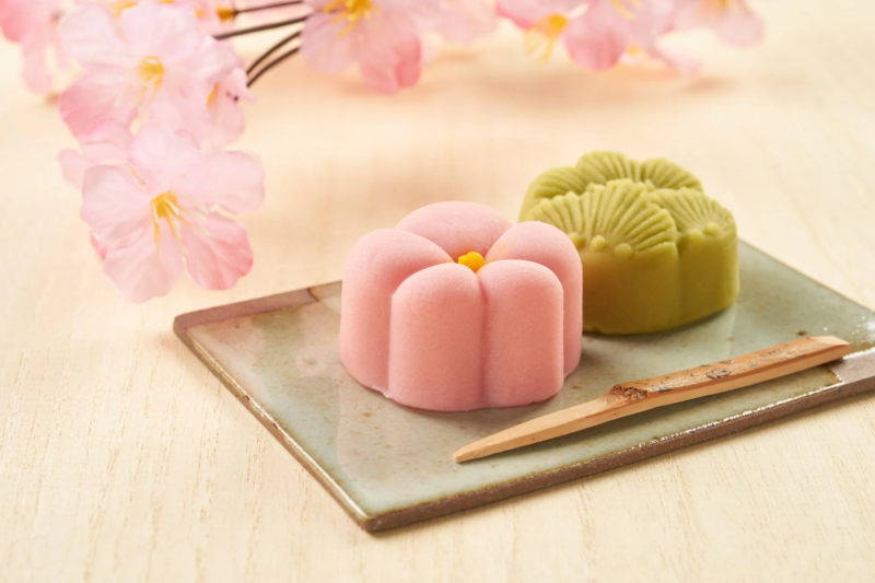 桜の花を背景に、ピンクと緑の美しい和菓子が皿に盛られています