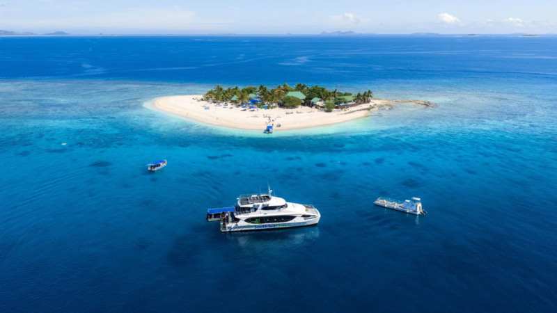 澄み切った青い海に浮かぶ熱帯の小さな島と白い砂浜が美しい空撮写真。島にはリゾート施設があり、周りにはクルーズ船や小型ボートが停泊し、観光客で賑わっている様子が伺えます。