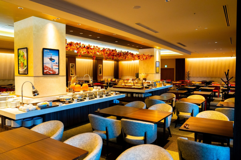 秋の装飾が施された広々としたビュッフェレストランの内部。温かい照明の下、料理カウンターにはカボチャが飾られ、多数のテーブルと椅子が配置されています。柱にはスキーとサラダのポスターが見えます。