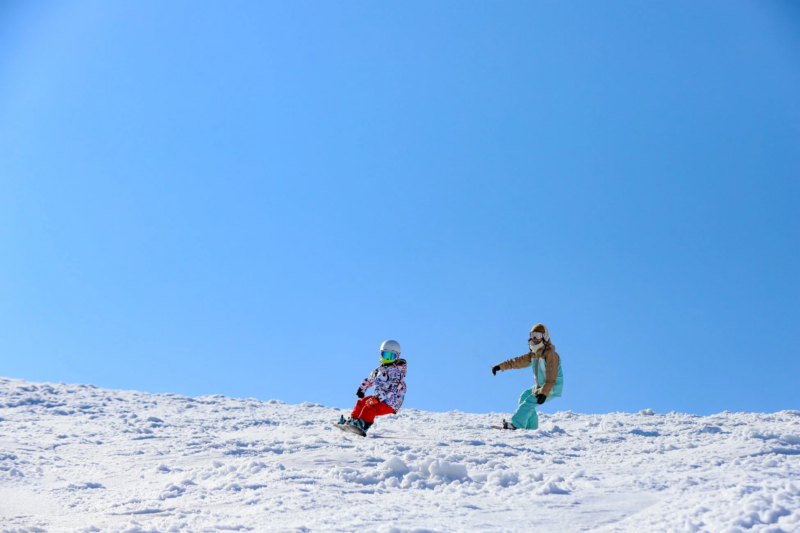 晴れた青空の下、雪山でスノーボードを楽しむ二人の姿を捉えた写真です。広々としたゲレンデを滑走しており、冬のレジャースポーツの魅力が伝わります。