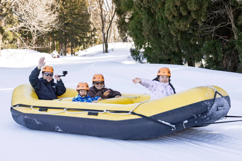 雪景色の広がる場所で、家族4人がインフレータブルボートに乗ってスノーラフティングを楽しんでいます。全員がオレンジ色のヘルメットを着用し、笑顔で手を振るなどして、冬のアクティビティを満喫している様子が伝わります。