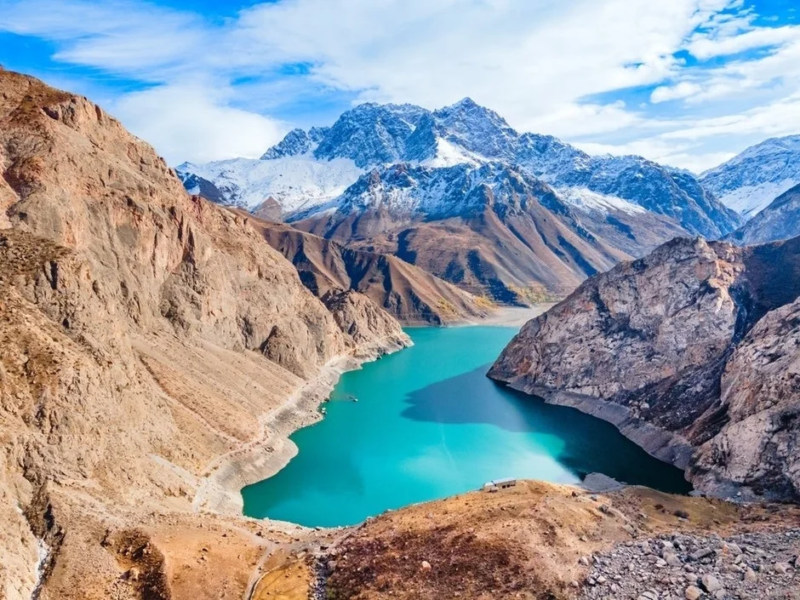 険しい岩山に囲まれた、鮮やかなターコイズブルーの湖が広がる壮大な自然景観です。遠くには雪をかぶった山々が連なり、青空の下、息をのむような美しい景色が広がっています。