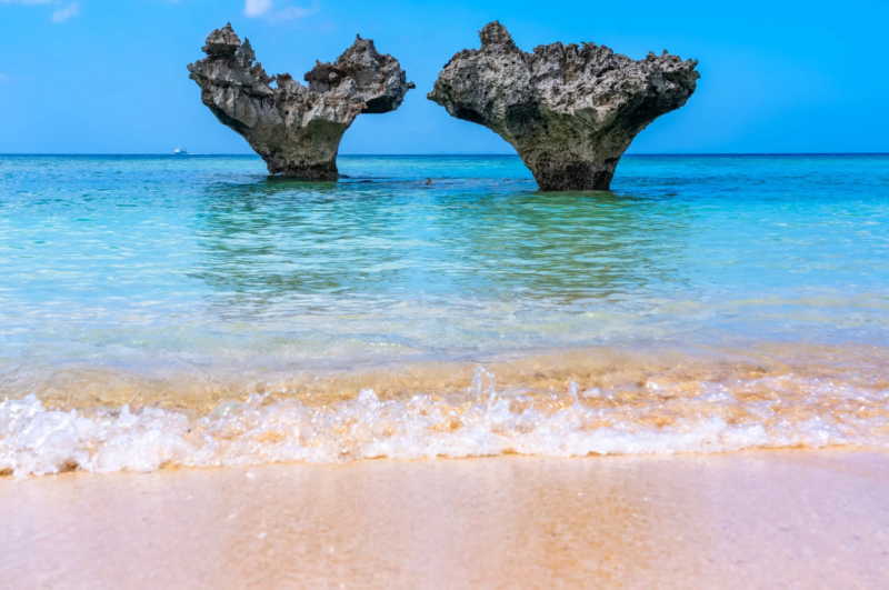 青い空と透明な海が広がるビーチに、ハートの形をした岩が二つ立っています。