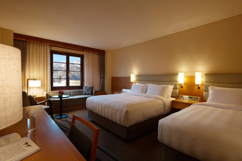 雪山が見える窓があるホテルのツインルームです。温かい照明の室内には、2つのベッド、ソファ、デスクが配置されており、快適で落ち着いた雰囲気が漂っています。