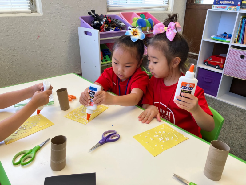 二人の幼い女の子がテーブルで工作活動に集中しています。彼女たちは赤いTシャツを着ており、のりを使って紙に模様を描いています。教室のような環境で、創造的な遊びを楽しんでいる様子がうかがえます。