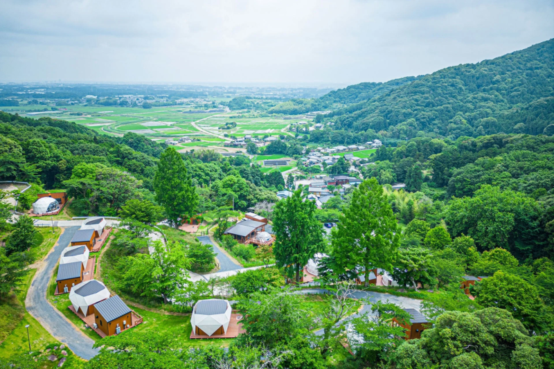 緑豊かな山腹にモダンなデザインの宿泊施設が点在し、その向こうには田園風景と集落が広がる雄大な景色