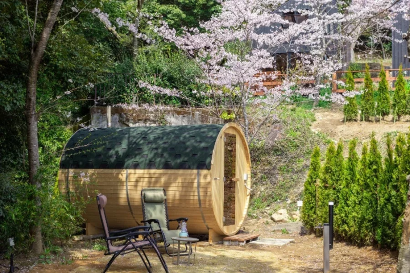 桜が咲き誇る自然豊かな場所に設置された、木製のバレルサウナと屋外の休憩スペース
