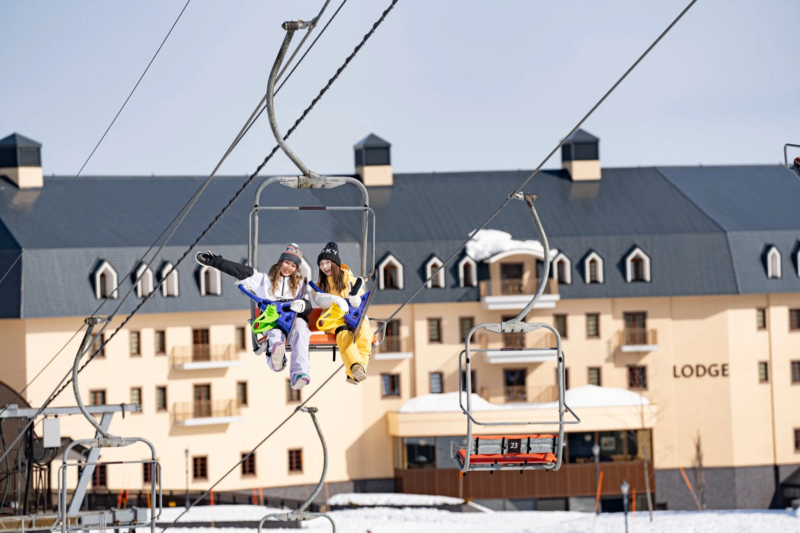 雪山でスキーリフトに乗る楽しそうな二人の女性。背景には大きなロッジが見え、冬のレジャーを満喫している様子が伝わります。