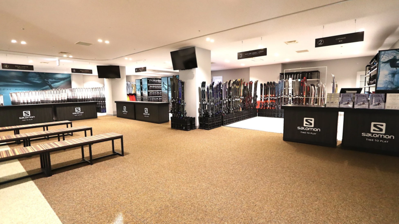 サロモンのスキー・スノーボード用品店の広々とした店内。多数のスキー板が展示され、レンタルカウンター、ハイエンドモデルコーナー、会計がある。顧客用のベンチも設置されており、冬のスポーツ用品が充実している。