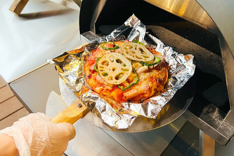 手袋をはめた手がピザピールを使って、蓮根やピーマンが乗ったピザをアルミホイルごとオーブンに入れようとしている様子