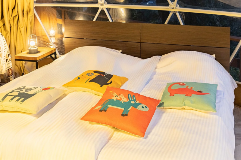 ドーム型テント内のベッドルームで、白い寝具と可愛い動物柄のカラフルなクッションが複数並べられています