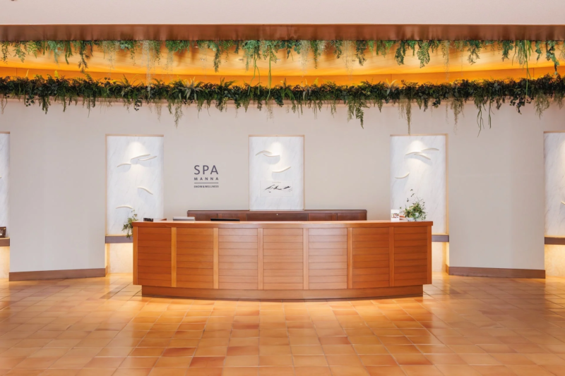 木製の受付カウンターと壁の装飾が特徴的な、明るく広々としたスパまたはホテルのロビーです。天井からは豊かな緑の植物が吊るされ、自然で落ち着いた雰囲気を作り出しています。