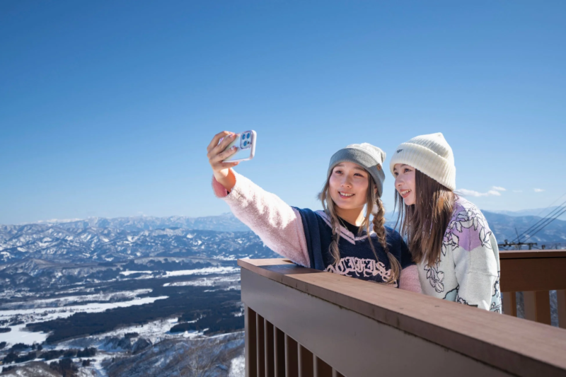 晴れた冬の日に、二人の女性が雪山の展望台から広大な雪景色を背景に自撮りを楽しんでいます。笑顔で思い出を作っている様子です。