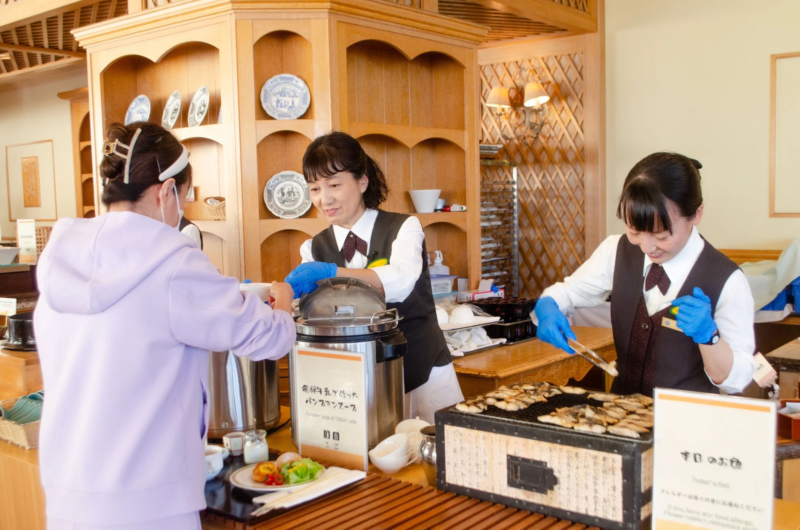 ホテルまたはレストランのビュッフェ朝食会場で、スタッフがゲストに焼き魚やパンプキンスープなどの温かい料理を提供している場面です。