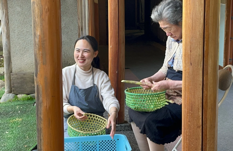 屋外で、笑顔の若い女性と年配の女性がタケノコの下処理をしている様子。
