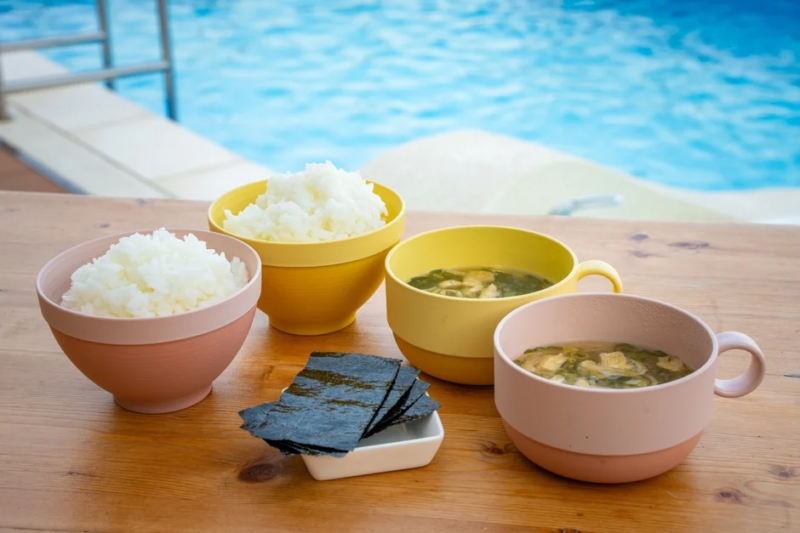 木製のテーブルに、ご飯、味噌汁、海苔の和食が並べられています。背景には青いプールの水が広がり、リゾートのような開放的な屋外での食事風景です。
